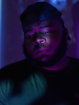 A thoughtful man in a cap stands under dramatic purple lighting indoors.
